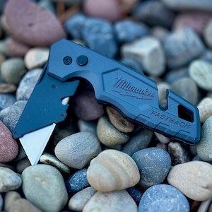 May include: A matte dark blue Milwaukee Fastback folding utility knife with a partially extended silver metal blade. The handle features the embossed brand name Milwaukee and the model name Fastback, with a small circular button near the pivot point.