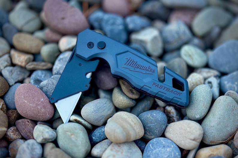 May include: A matte dark blue Milwaukee Fastback folding utility knife with a partially extended silver metal blade. The handle features the embossed brand name Milwaukee and the model name Fastback, with a small circular button near the pivot point.