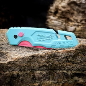 May include: A light blue Milwaukee Fastback utility knife featuring a bright pink release button and a partially visible pink blade edge. The tool has a textured handle with the brand name and model name embossed on the side, and a lanyard hole.