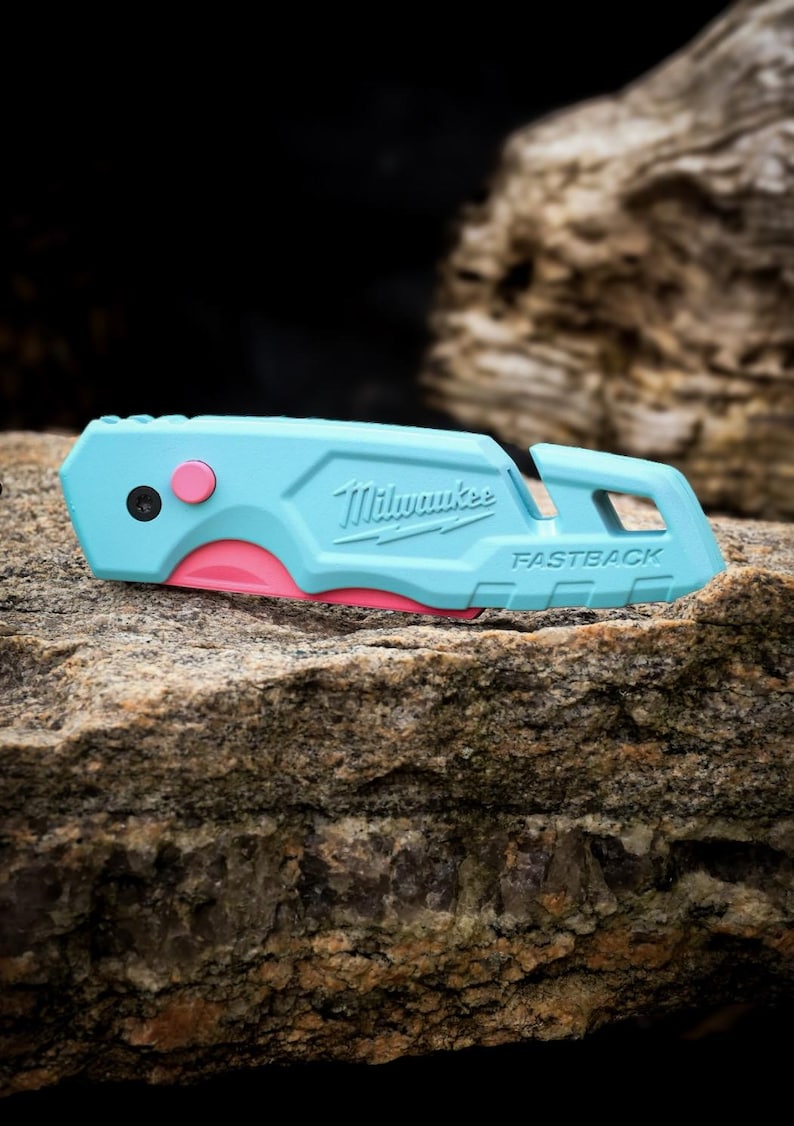 May include: A light blue Milwaukee Fastback utility knife featuring a bright pink release button and a partially visible pink blade edge. The tool has a textured handle with the brand name and model name embossed on the side, and a lanyard hole.