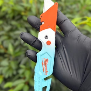 May include: A Milwaukee Fastback utility knife featuring a folding metal blade and a handle with a color gradient transitioning from bright orange at the top to white in the middle and light blue at the bottom, with the brand name printed in red.