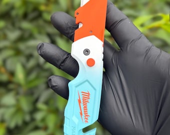 以下が含まれることがあります： 折りたたみ式の金属製ブレードを備えたMilwaukee Fastbackユーティリティナイフ。ハンドルは上部が鮮やかなオレンジ、中央が白、下部がライトブルーのグラデーションになっており、赤い文字でブランド名がプリントされています。