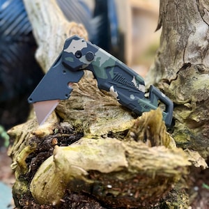 May include: A folding utility knife with a camouflage pattern handle featuring the Milwaukee brand name and the word FASTBACK embossed on the side. The tool has a matte black metal blade holder and a partially extended silver utility blade.