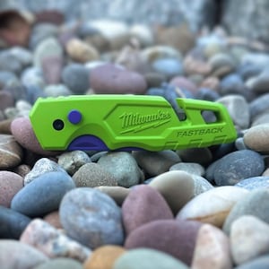 May include: A lime green Milwaukee Fastback utility knife featuring a purple button and a purple accent near the blade area. The brand name Milwaukee and the word Fastback are embossed on the side of the handle, which has a textured grip.