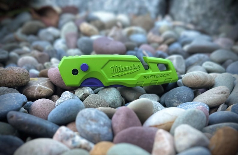 May include: A lime green Milwaukee Fastback utility knife featuring a purple button and a purple accent near the blade area. The brand name Milwaukee and the word Fastback are embossed on the side of the handle, which has a textured grip.