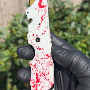 May include: A utility knife featuring a white handle with a red splatter pattern and a gold-coloured blade. The handle has a textured grip, a central circular button, and a small hole at the base for hanging or attachment purposes.
