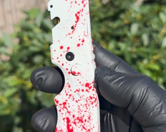 以下が含まれることがあります： 赤色のスプラッター模様が施された白いハンドルと、金色の刃を持つユーティリティナイフ。ハンドルにはテクスチャ加工が施されており、中央に円形のボタン、底部には吊り下げや固定用の小さな穴が開いています。