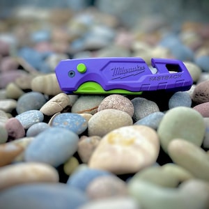 May include: A purple Milwaukee Fastback utility knife with a bright lime green blade and a small lime green button on the side. The tool features the Milwaukee logo and the word Fastback embossed on the side of the purple handle casing.