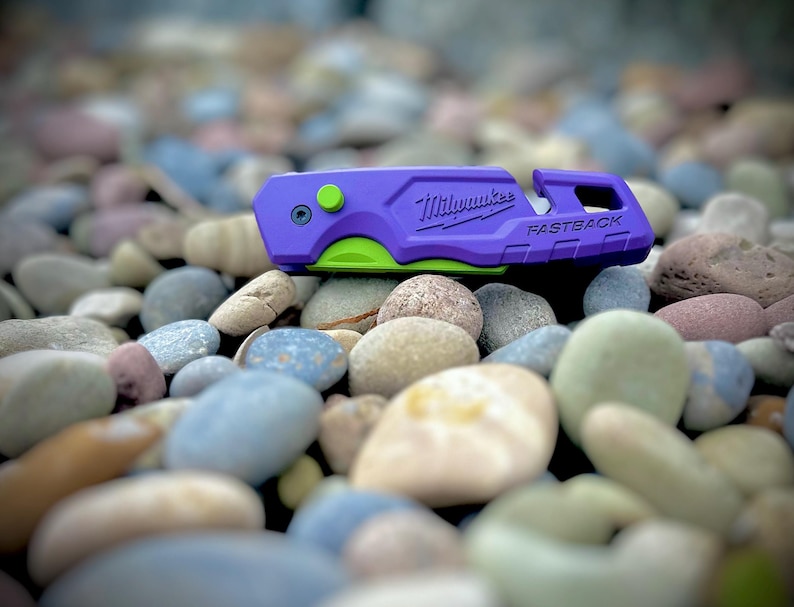 May include: A purple Milwaukee Fastback utility knife with a bright lime green blade and a small lime green button on the side. The tool features the Milwaukee logo and the word Fastback embossed on the side of the purple handle casing.
