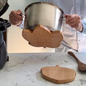 Könnte beinhalten: Ein heller Holzuntersetzer in Form eines Ahornblatts mit der Gravur CRAFTIVA, der einen Edelstahltopf stützt. Ein zweiter Untersetzer in Eichelform liegt auf der darunter liegenden Fläche, zusammen mit einem Holzlöffel.