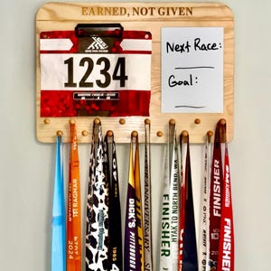 May include: A light-colored wooden wall-mounted display rack with the text "EARNED, NOT GIVEN" engraved at the top. It features a clip for a race bib numbered 1234, a whiteboard section labeled "Next Race:" and "Goal:", and pegs holding multiple colorful race medals.