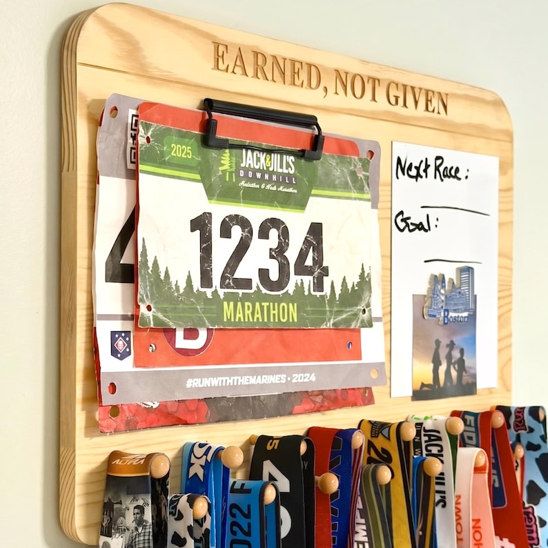 K&ouml;nnte beinhalten: Ein helles Holz-Displayboard mit der Gravur EARNED, NOT GIVEN. Es verf&uuml;gt &uuml;ber einen schwarzen Clip f&uuml;r Startnummern, ein wei&szlig;es Whiteboard-Feld mit der Beschriftung Next Race und Goal sowie mehrere Holzhaken zum Aufh&auml;ngen von Medaillen.
