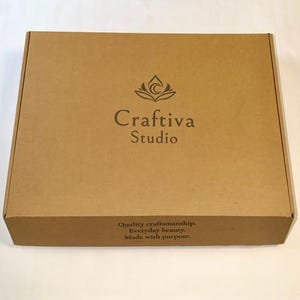 K&ouml;nnte beinhalten: Ein rechteckiger brauner Karton mit dem dunkelbraunen Logo und dem Schriftzug Craftiva Studio auf dem Deckel. Auf der Vorderseite des Kartons steht der Text Quality craftsmanship. Everyday beauty. Made with purpose. in dunkelbrauner Schrift.