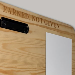 K&ouml;nnte beinhalten: Ein helles Holz-Klemmbrett mit einer schwarzen Metallklemme auf der linken Seite. Die Holzmaserung ist auf der gesamten Oberfl&auml;che sichtbar. Der Schriftzug EARNED, NOT GIVEN ist in Gro&szlig;buchstaben am oberen Rand des Brettes eingraviert.