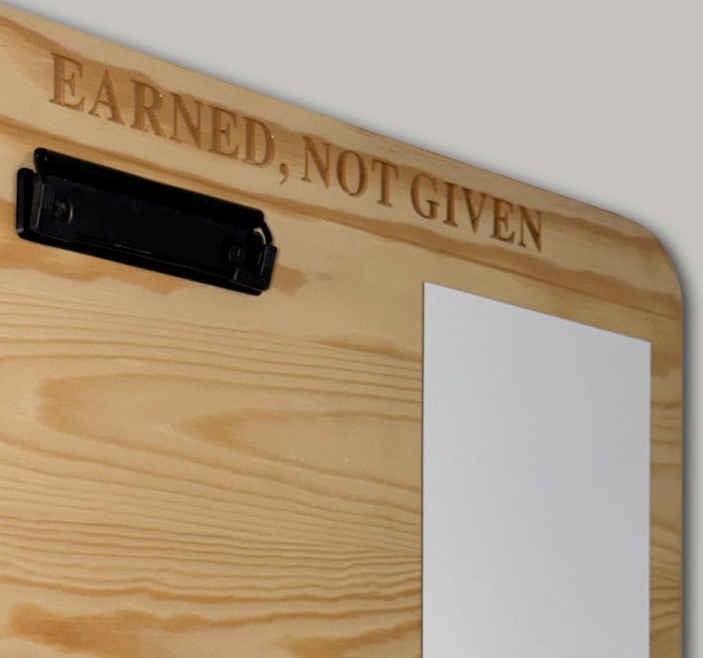 K&ouml;nnte beinhalten: Ein helles Holz-Klemmbrett mit einer schwarzen Metallklemme auf der linken Seite. Die Holzmaserung ist auf der gesamten Oberfl&auml;che sichtbar. Der Schriftzug EARNED, NOT GIVEN ist in Gro&szlig;buchstaben am oberen Rand des Brettes eingraviert.