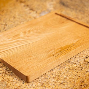 Könnte beinhalten: Ein rechteckiges, hellbraunes Holzschneidebrett mit sichtbarer natürlicher Holzmaserung. Das Brett verfügt über eine horizontale, eingelassene Rille nahe der Oberkante und das Wort CRAFTIVA, das in einfacher Schrift unten rechts eingraviert ist.