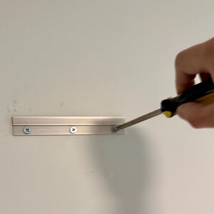 K&ouml;nnte beinhalten: Eine silberfarbene Wandmontagehalterung aus Metall mit horizontalem Profil, die drei vorgebohrte L&ouml;cher aufweist und mit silbernen Schrauben an einer flachen Oberfl&auml;che befestigt ist, w&auml;hrend ein Schraubendreher die rechte Schraube festzieht.