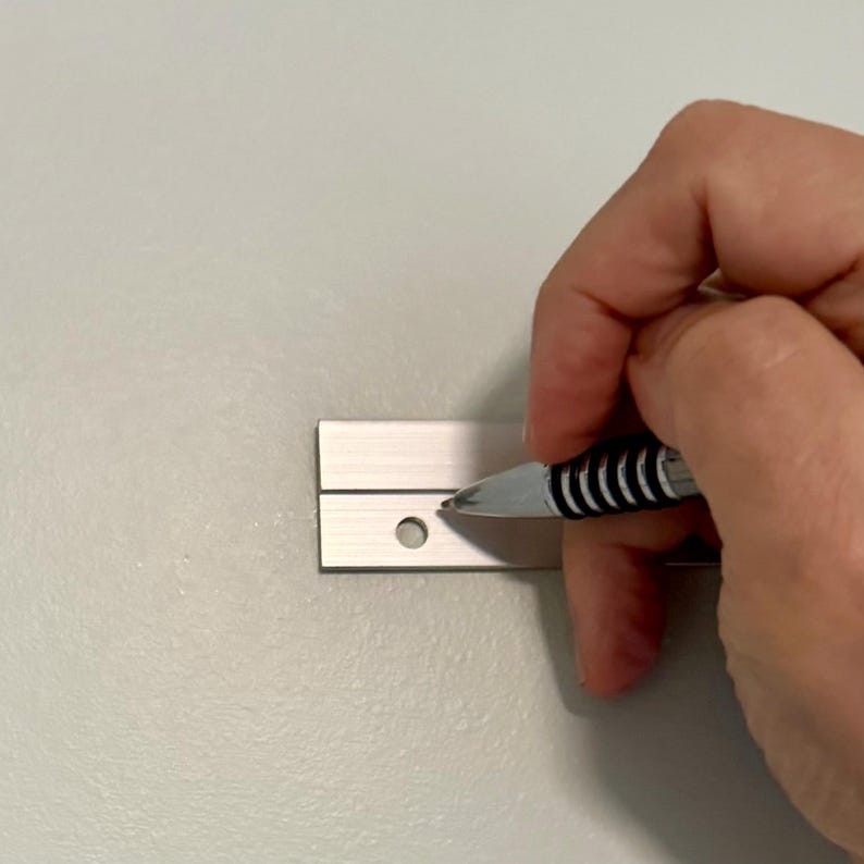 K&ouml;nnte beinhalten: Ein silberfarbener Metallstreifen mit einer horizontalen Nut und einem kreisf&ouml;rmigen Loch, der horizontal ausgerichtet ist. Das Metallteil hat eine glatte, geb&uuml;rstete Oberfl&auml;che und eine rechteckige Form mit einem vorgebohrten Loch.