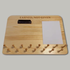 K&ouml;nnte beinhalten: Ein rechteckiges Display-Board aus hellem Holz mit einer schwarzen Metallklemme oben, dem eingravierten Text "EARNED, NOT GIVEN" und einem leeren wei&szlig;en rechteckigen Blatt Papier. Der untere Bereich hat zwei Reihen kleiner Holzstifte zum Aufh&auml;ngen.