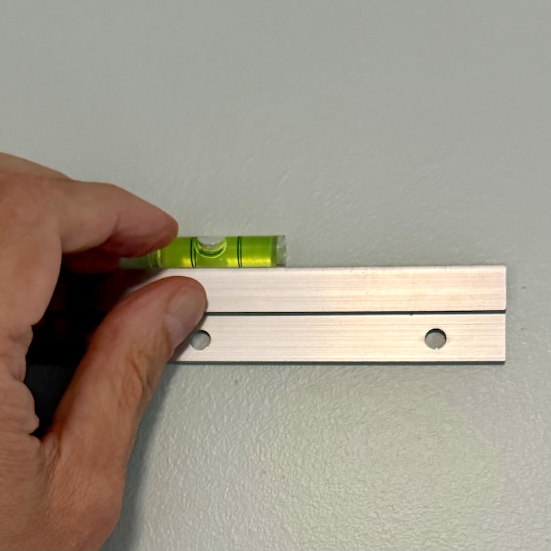 K&ouml;nnte beinhalten: Eine silberfarbene Z-Profil-Wandhalterung aus Metall mit zwei vorgebohrten runden Montagel&ouml;chern, die horizontal an einer Wand angebracht ist, wobei eine kleine gr&uuml;ne Wasserwaage auf der Oberkante liegt, um eine gerade Ausrichtung zu gew&auml;hrleisten.