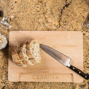 Könnte beinhalten: Ein rechteckiges, helles Holzschneidebrett mit dem eingravierten Wort CRAFTIVA am unteren Rand. Ein Edelstahl-Kochmesser mit schwarzem Griff liegt auf dem Brett neben mehreren Scheiben von rustikalem, krustigem Brot.