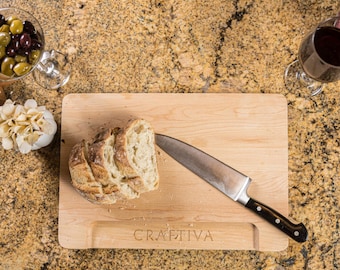 Könnte beinhalten: Ein rechteckiges, helles Holzschneidebrett mit dem eingravierten Wort CRAFTIVA am unteren Rand. Ein Edelstahl-Kochmesser mit schwarzem Griff liegt auf dem Brett neben mehreren Scheiben von rustikalem, krustigem Brot.