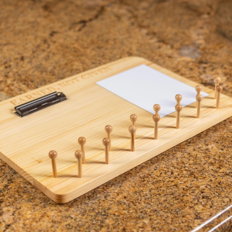 K&ouml;nnte beinhalten: Ein rechteckiges Display-Board aus hellem Holz mit einem schwarzen Metallclip auf der linken Seite und dem eingravierten Text EARNED, NOT GIVEN. Ein wei&szlig;es quadratisches Papier ist auf dem Board befestigt, das zehn vertikale Holzstifte enth&auml;lt.