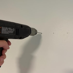 K&ouml;nnte beinhalten: Eine schwarze elektrische Bohrmaschine mit Metallbohrfutter und Bohrer, die an einer schlichten wei&szlig;en Wand angesetzt ist. Das Geh&auml;use der Bohrmaschine zeigt aufgedruckte technische Spezifikationen und Sicherheitshinweise, einschlie&szlig;lich Text zu 120 Volt.