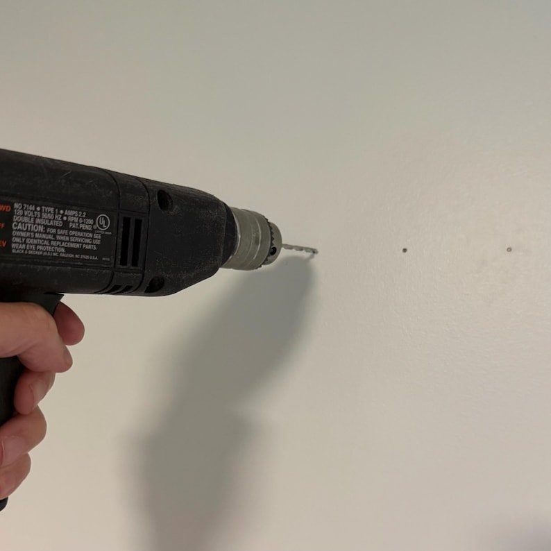 K&ouml;nnte beinhalten: Eine schwarze elektrische Bohrmaschine mit Metallbohrfutter und Bohrer, die an einer schlichten wei&szlig;en Wand angesetzt ist. Das Geh&auml;use der Bohrmaschine zeigt aufgedruckte technische Spezifikationen und Sicherheitshinweise, einschlie&szlig;lich Text zu 120 Volt.