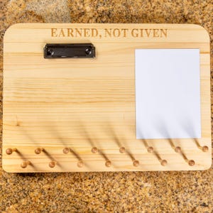 K&ouml;nnte beinhalten: Ein rechteckiges Display-Board aus hellem Holz mit dem eingravierten Text EARNED, NOT GIVEN. Es enth&auml;lt eine schwarze Metallklemme oben links, ein leeres wei&szlig;es rechteckiges Blatt Papier auf der rechten Seite und sechzehn kleine Holzstifte.