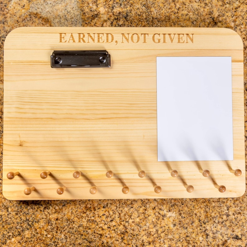 K&ouml;nnte beinhalten: Ein rechteckiges Display-Board aus hellem Holz mit dem eingravierten Text EARNED, NOT GIVEN. Es enth&auml;lt eine schwarze Metallklemme oben links, ein leeres wei&szlig;es rechteckiges Blatt Papier auf der rechten Seite und sechzehn kleine Holzstifte.