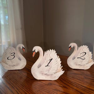 May include: Three white swan-shaped table markers made of wood, each featuring a hand-painted orange beak and black eyes. The swans are numbered one, two, and three in black script on their wings, designed to stand upright on a flat surface.
