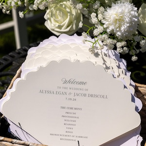 Könnte beinhalten: Ein Stapel cremefarbener Hochzeitsfächer mit gewelltem Rand in einem geflochtenen Korb. Der Text lautet: Welcome to the wedding of Alyssa Egan & Jacob Driscoll 7.19.26, gefolgt vom Ablauf der Zeremonie: Prelude, Processional, Welcome, Readings, Vows, Rings, Pronouncement of Marriage, Recessional.