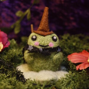 May include: A whimsical felted frog wearing a brown witch hat and a black collar with star accents. The frog has pink cheeks and sits on a cloud-like base, surrounded by flowers and moss.