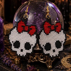 May include: A pair of beaded earrings shaped like white skulls with black eye sockets and a red bow on top. The earrings feature gold-toned metal hooks and are crafted from small, uniform beads arranged in a grid pattern to form the skull design.