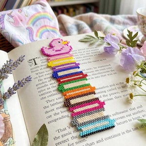 Könnte beinhalten: Ein Perlen-Lesezeichen, das einen Stapel bunter Bücher mit einer kleinen rosa Ente an der Spitze darstellt. Das Lesezeichen besteht aus winzigen, gleichmäßigen Perlen in Gelb, Blau, Rot, Grün, Orange, Violett, Grau und Hellblau in Reihen.