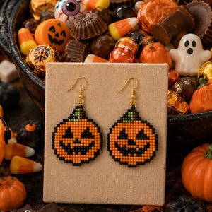 May include: A pair of beaded dangle earrings featuring a pixelated jack-o'-lantern pumpkin design in orange, black, and green. The earrings have gold-toned metal hooks and are displayed on a rectangular beige card against a Halloween candy background.