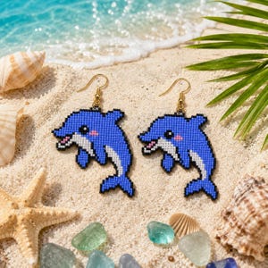 Könnte beinhalten: Ein Paar Ohrhänger mit einem gepixelten Delfin-Design aus kleinen blauen und weißen Perlen. Jeder Delfin hat einen weißen Bauch, ein rosa Wangendetail und einen Ohrhaken aus goldfarbenem Metall zur sicheren Befestigung am Ohr.