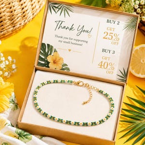 May include: A beaded anklet featuring a repeating pattern of small white and yellow daisy flowers on a green background, finished with a gold-tone metal lobster clasp and extender chain, presented inside a cardboard box with a thank you card.
