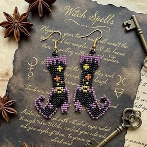 May include: A pair of beaded earrings shaped like curled-toe boots. The boots are primarily black with purple cuffs and heels, accented with small yellow and orange floral-like bead patterns. Each earring features a gold-toned metal hook.