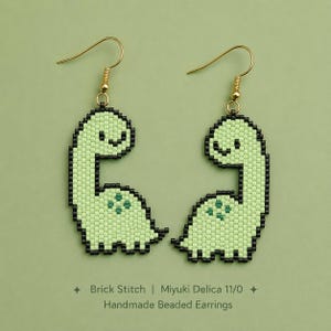 May include: A pair of handmade beaded earrings featuring a light green dinosaur design with black outlines and gold-tone hooks. The text below reads: Brick Stitch | Miyuki Delica 11/0 | Handmade Beaded Earrings. The beads are arranged in a pixelated style.