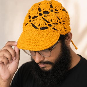 Puede incluir: Una gorra de béisbol de ganchillo color amarillo mostaza con un intrincado patrón calado de inspiración floral en la copa y una visera sólida de punto apretado. La gorra incluye un fino cordón de ganchillo a juego en la parte trasera.