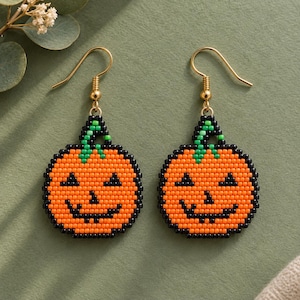 Puede incluir: Un par de pendientes colgantes de cuentas con un diseño de calabaza de Halloween. Las calabazas están hechas de cuentas de semillas naranjas, negras y verdes, mostrando una cara sonriente con ojos triangulares y un tallo verde, con ganchos dorados.