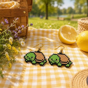 Peut inclure: Une paire de boucles d'oreilles pendantes représentant des tortues pixelisées en perles. Les tortues ont une tête vert clair, une carapace beige et des contours noirs, suspendues à des crochets en métal doré pour oreilles percées.