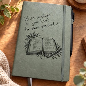 May include: A sage green notebook with an elastic closure and a black pen. The cover features the text "Write scripture on your heart for when you need it" above a detailed black line drawing of an open Bible surrounded by leafy branches.