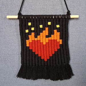 Op de afbeelding: Zwarte macramé wandhanger met een gepixeld hartontwerp in rood, oranje en geel. De wandhanger hangt aan een houten stok en heeft een franje aan de onderkant. De achtergrond is een lichtblauwe stof.