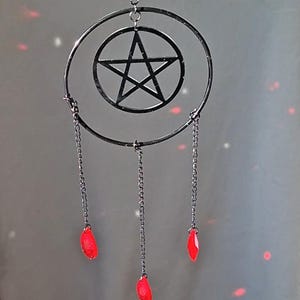 May include: A black metal pentagram suncatcher with a circular frame and three dangling chains. Each chain ends with a red, teardrop-shaped crystal. The suncatcher is designed to hang and catch light.