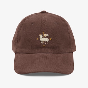 Peut inclure: Une casquette de baseball en velours côtelé marron foncé avec une broderie sur le devant. La broderie représente un agneau blanc debout à côté d'une grande croix avec un drapeau, entouré d'une petite étoile dorée et d'un soleil doré.