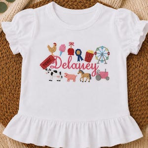 May include: White ruffle-sleeve t-shirt with a colorful embroidered design. The design includes a chicken, cotton candy, a ribbon, popcorn, a Ferris wheel, and a tractor. The name "Delaney" is also embroidered on the shirt.