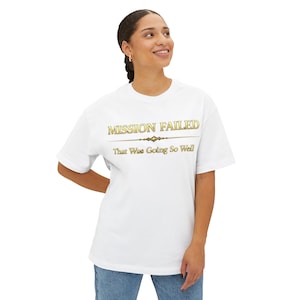 Könnte beinhalten: Weißes T-Shirt mit goldfarbenem Schriftzug „MISSION FAILED“ über „That Was Going So Well“. Das kurzärmlige T-Shirt hat eine lockere Passform und eignet sich für Freizeitkleidung.